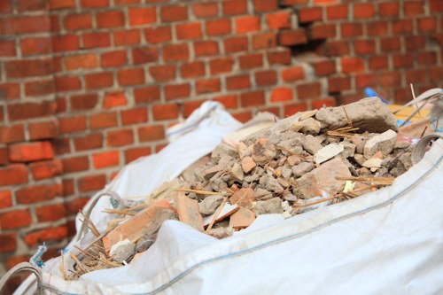 A large white sack filled with a mixture of construction debris including broken bricks, concrete fragments, and small pieces of tiles, positioned outdoors on a paved surface in front of a red brick wall. The debris appears to be recently collected and contains a variety of textures and earthy tones such as reddish-brown, beige, and gray. The sackâ€™s material is crinkled and slightly translucent, revealing the contents inside. To the left, a glimpse of a metal object, possibly part of waste management equipment, is visible. The scene suggests a punctual clearance of building waste, consistent with services offered by Office Clearance Sanderstead, operating in the South London area near Sanderstead, highlighting the process of rubbish removal in a residential or commercial setting.