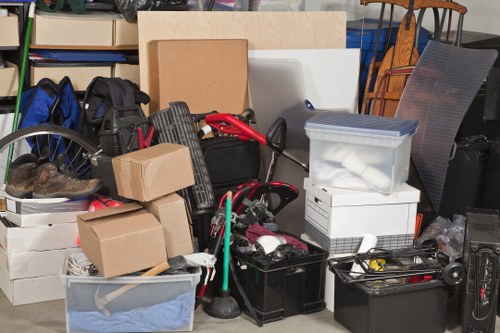 A cluttered indoor space filled with various household and office items prepared for rubbish removal, including cardboard boxes of different sizes, some sealed and others open revealing clothing and soft items, a clear plastic storage box with a blue lid, a black plastic toolbox, and a green-handled mop resting among the debris. Other visible objects include a bicycle wheel, a black backpack, and a variety of miscellaneous tools and equipment. The items are stacked and arranged in a somewhat compact manner, with some leaning against each other or placed atop one another, occupying a concrete or tiled floor area typical of a garage or storage room. The background features shelving units and a chair, indicating a domestic or commercial environment. The scene is well-lit, with natural and artificial light highlighting the textures of cardboard, plastic, metal, and fabric surfaces, consistent with a waste clearance operation such as those undertaken by Office Clearance Sanderstead in the local Sanderstead area, near Croydon.
