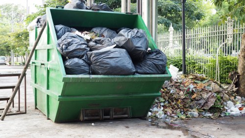 A large green skip bin situated outdoors on a paved area near a grass verge with a metal fence and trees in the background. The bin is filled with tightly packed black plastic rubbish bags, overflowing slightly at the top. To the right of the skip, there is a small pile of mixed waste including cardboard, paper, and plastic debris on the ground. The environment appears to be a residential or park area, with natural light illuminating the scene, emphasizing the materials' textures and the organized but cluttered state of the rubbish ready for removal, consistent with waste disposal services in Sanderstead or nearby South London locations. The scene is captured in a straightforward, factual manner, reflecting typical rubbish clearance activity that Office Clearance Sanderstead might handle.