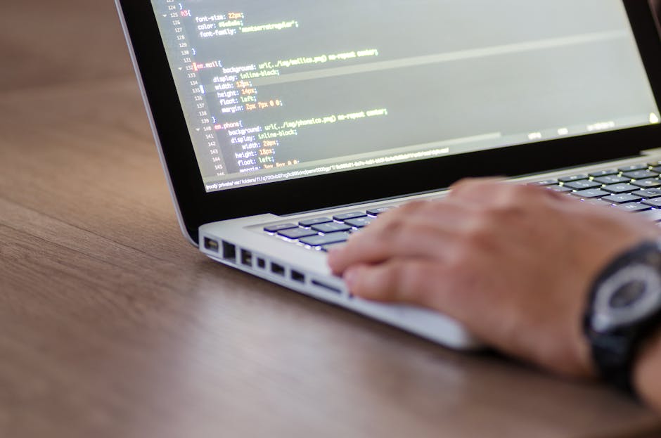 A person with a beard and glasses, wearing a grey sweater, is seated at a wooden desk working on a computer with dual monitors. The left monitor displays a directory or file management interface in da