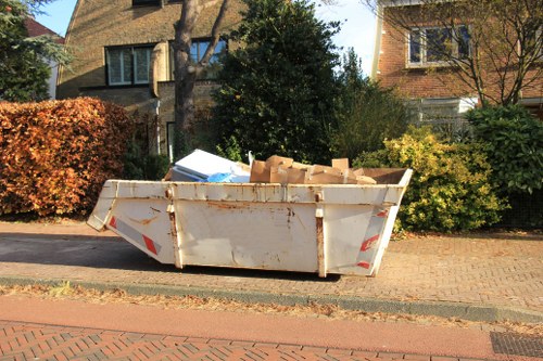 A large, white metal skip container filled with scattered cardboard boxes and packaging material, positioned on a paved residential street with a brick pavement. The skip, exhibiting signs of rust and weathering, is situated in front of a garden area with shrubs and trees, behind which there are two detached houses with window details and brick or stone facades. Sunlight illuminates the scene, casting shadows and highlighting the materials and textures of the skip and surrounding environment. The image suggests a rubbish removal service, possibly by Office Clearance Sanderstead, operating within the local area near Sanderstead postcode.